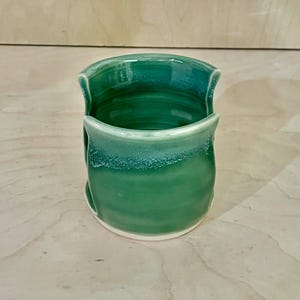 May include: A cylindrical ceramic sponge holder with a glossy emerald green glaze. The vessel features a wide, U-shaped cutout on the front and back, revealing the interior. The rim and base show subtle variations in color intensity and texture.