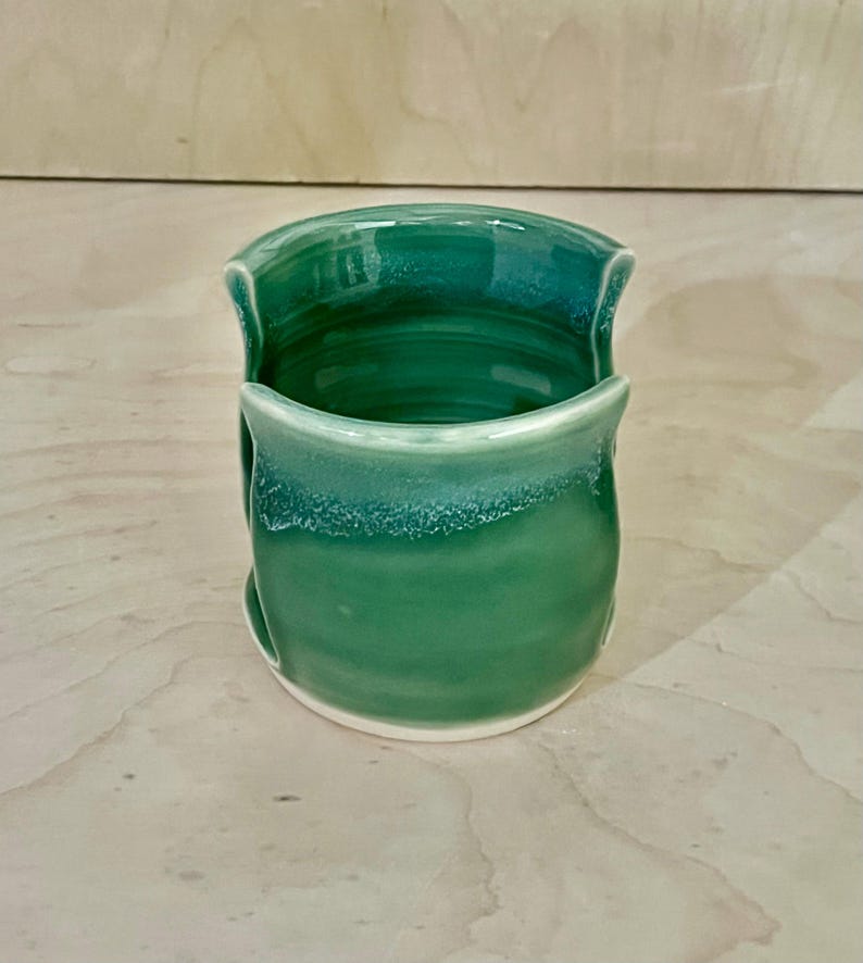 May include: A cylindrical ceramic sponge holder with a glossy emerald green glaze. The vessel features a wide, U-shaped cutout on the front and back, revealing the interior. The rim and base show subtle variations in color intensity and texture.