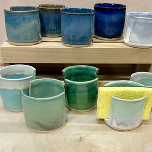 May include: A collection of ten ceramic sponge holders in various shades of blue, green, and white. Each holder features a U-shaped cutout on the front rim. One holder contains a rectangular yellow sponge. The items are arranged on two levels.