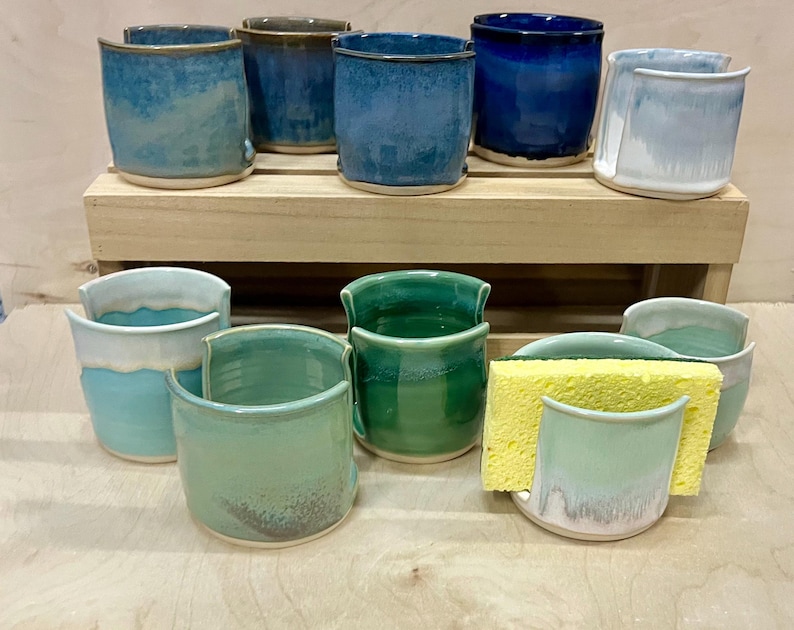May include: A collection of ten ceramic sponge holders in various shades of blue, green, and white. Each holder features a U-shaped cutout on the front rim. One holder contains a rectangular yellow sponge. The items are arranged on two levels.