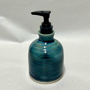 Pu&ograve; includere: Un dispenser di sapone in ceramica con una smaltatura lucida blu e verde acqua, caratterizzato da texture a coste orizzontali sul corpo. Il contenitore ha una forma arrotondata ed &egrave; dotato di una pompa in plastica nera funzionale.