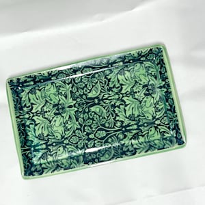 May include: A rectangular ceramic tray featuring a dark green and light green botanical pattern. The design includes intricate illustrations of rabbits, birds, and dense foliage. The tray has a smooth, glossy finish and a light green border.