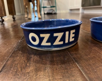 May include: A deep blue ceramic pet bowl with a glossy finish featuring the name OZZIE embossed in large white capital letters on the front side. The bowl has a wide circular opening and a thick rim, with a second matching bowl partially visible.