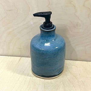 Pu&ograve; includere: Un dispenser per sapone in ceramica con corpo arrotondato caratterizzato da una smaltatura blu screziata che sfuma in una tonalit&agrave; pi&ugrave; scura verso la base. Il dispenser presenta una pompa in plastica nera e un bordo bianco visibile sul fondo.