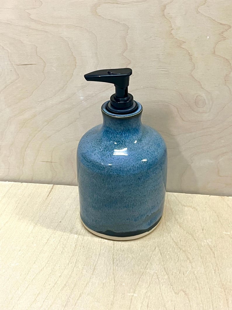Pu&ograve; includere: Un dispenser per sapone in ceramica con corpo arrotondato caratterizzato da una smaltatura blu screziata che sfuma in una tonalit&agrave; pi&ugrave; scura verso la base. Il dispenser presenta una pompa in plastica nera e un bordo bianco visibile sul fondo.