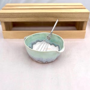 May include: A handmade ceramic mixing bowl with a light green and blue glaze, containing a white substance, and a stainless steel whisk. The bowl has a small handle and a pouring spout. A wooden crate is in the background.