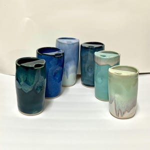 May include: A collection of six cylindrical ceramic tumblers with integrated lids, featuring a variety of glossy glaze finishes in shades of deep teal, royal blue, light sky blue, dark navy, turquoise, and a pale mint green with white drip accents.