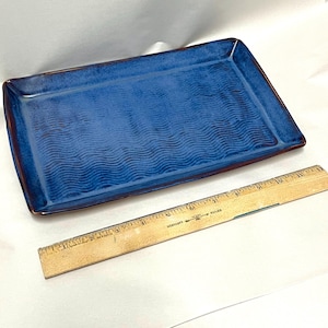 May include: A rectangular ceramic tray with a deep blue glaze and a textured wavy pattern on the surface. The tray features dark brown edges and is positioned next to a wooden ruler that shows the tray is approximately 11 inches in length.