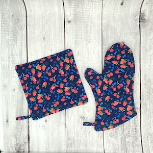 May include: A set of two blue pot holders with a pattern of red strawberries and blue blueberries. The pot holders are made of fabric and have a hanging loop.