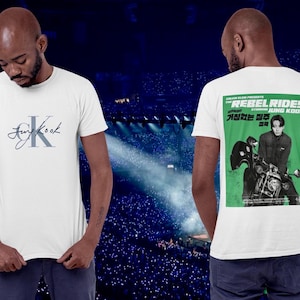 Peut inclure: T-shirt blanc avec le nom "Jungkook" en écriture manuscrite et un "K" stylisé sur le devant. Le dos du t-shirt présente un motif d'affiche verte avec le texte "REBEL RIDES" et une image d'une personne sur une moto.