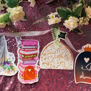 Puede incluir: Una guirnalda decorativa con varias formas recortadas, incluyendo un árbol, una figura con tela fluida, una pila de libros, una cúpula y una forma con una corona. La guirnalda está decorada con elementos florales y texto árabe, sobre un fondo rosa brillante.