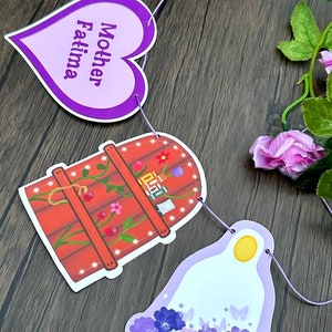 Puede incluir: Bandera de papel morado y blanco con el texto "Mother Fatima" en una pieza en forma de corazón, un pozo de madera marrón con flores y luces, y una campana morada con flores y mariposas.