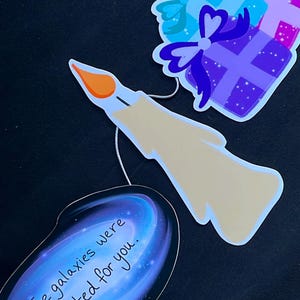 Puede incluir: Un conjunto de tres recortes de papel para una fiesta de cumpleaños. Los recortes son una vela, una galaxia con el texto "The galaxies were created for you." y una pila de regalos con purpurina morada, azul y rosa.