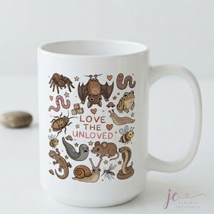 May include: A white ceramic mug featuring a colourful illustration of various small animals and insects, including a bat, spider, snake, and snail. The text "LOVE THE UNLOVED" is printed in the centre, surrounded by small hearts and stars.