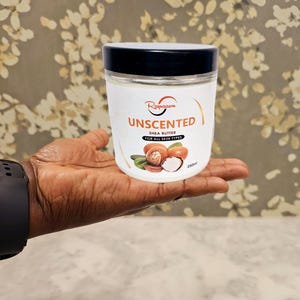 Op de afbeelding: Een witte pot met een zwarte deksel en een label met de tekst "Raynaasou Unscented Shea Butter for all skin types 250ml". De pot wordt in een hand gehouden.