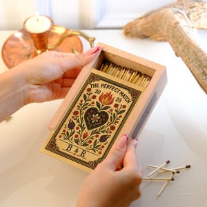 May include: A wooden matchbox with a decorative floral design and the text "THE PERFECT MATCH 20 25" is held open. The design includes a heart, flowers, and the initials "B & R". The matchbox contains numerous wooden matches.