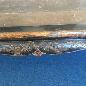 Può includere: Primo piano del bordo inferiore di un tavolo in legno nero con dettagli intagliati intricati. Il tavolo ha un piano di colore marrone scuro e una superficie blu sottostante.