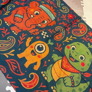 May include: A dark teal fabric with a colorful pattern featuring cartoon elephants, monsters, and turtles. The design includes paisley shapes, floral elements, and a checkered flag. The characters are in orange, yellow, and green.