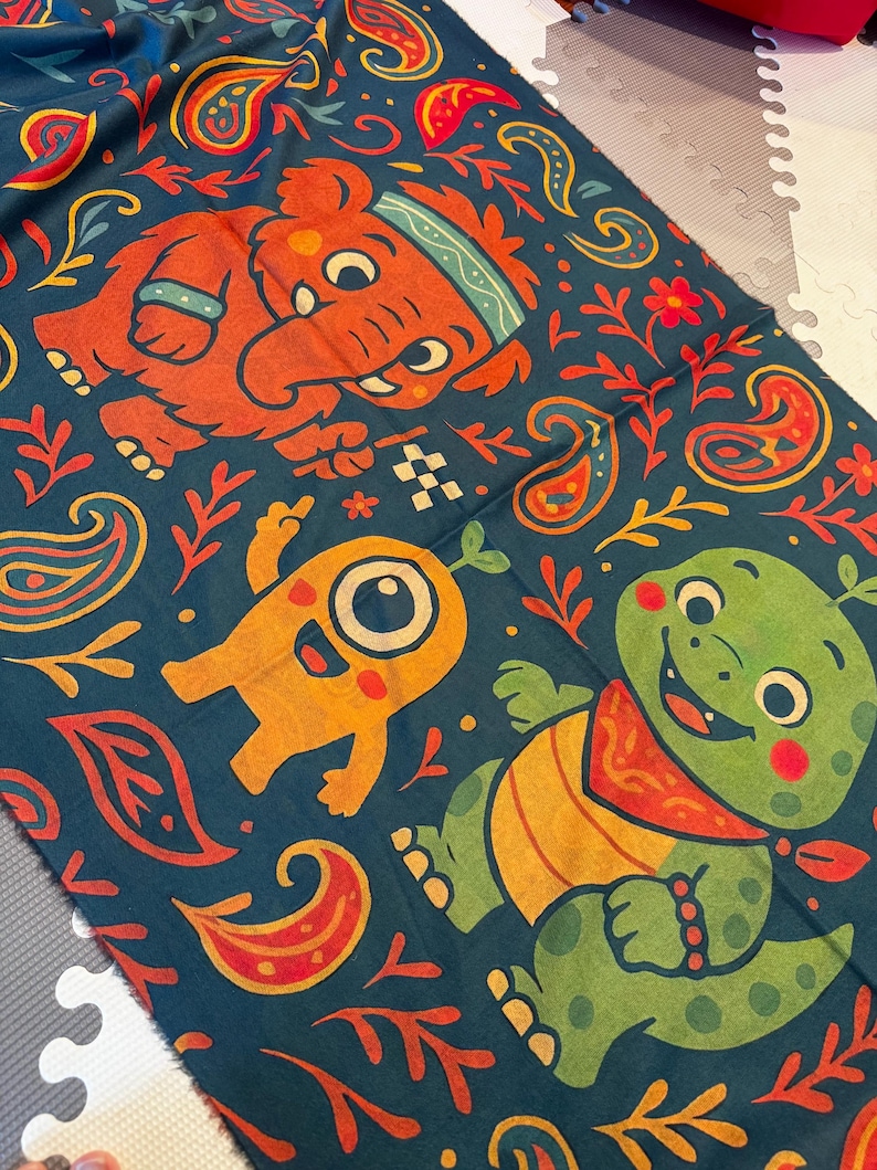 May include: A dark teal fabric with a colorful pattern featuring cartoon elephants, monsters, and turtles. The design includes paisley shapes, floral elements, and a checkered flag. The characters are in orange, yellow, and green.