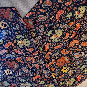 May include: Dark blue fabric with a repeating pattern of paisley shapes, cartoon bears, and smiling creatures in orange, red, yellow, and green. The fabric is folded, showing the pattern's continuity.