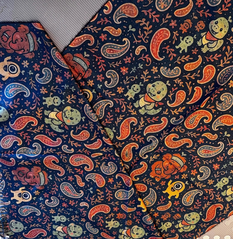 May include: Dark blue fabric with a repeating pattern of paisley shapes, cartoon bears, and smiling creatures in orange, red, yellow, and green. The fabric is folded, showing the pattern's continuity.