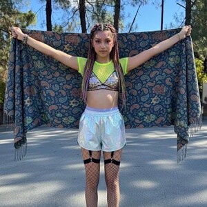 May include: A person holding a paisley patterned shawl with fringe. The person is wearing a neon green mesh top, a sequined bra, iridescent shorts, fishnet stockings, and black boots. The person has braided hair with pink and purple highlights.
