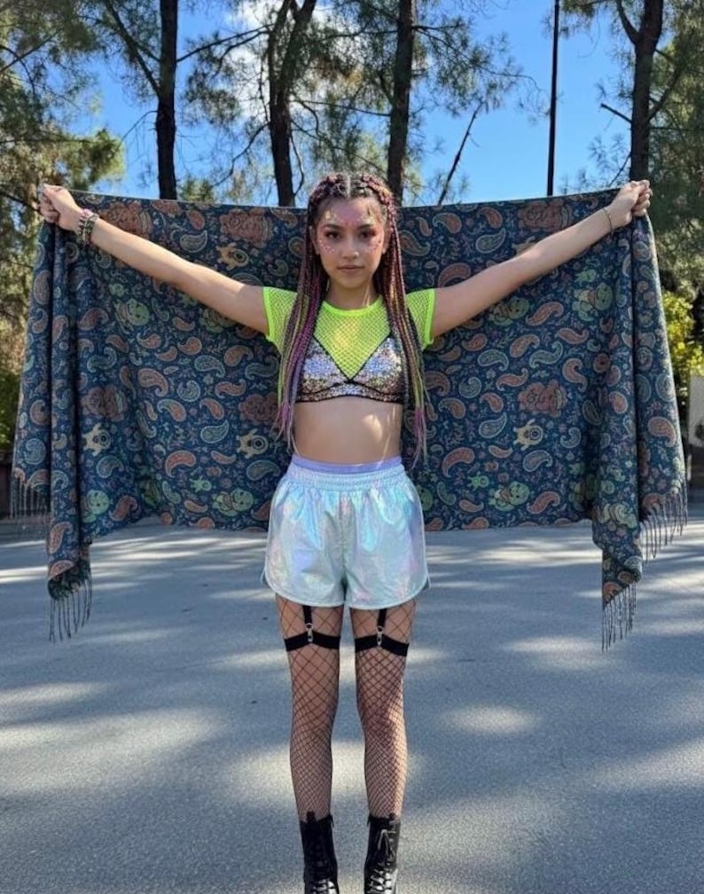 May include: A person holding a paisley patterned shawl with fringe. The person is wearing a neon green mesh top, a sequined bra, iridescent shorts, fishnet stockings, and black boots. The person has braided hair with pink and purple highlights.