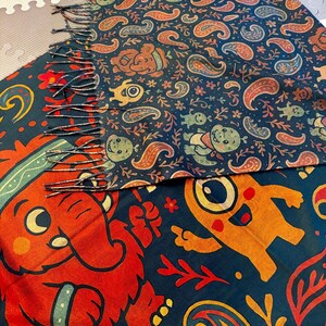 May include: A colorful scarf with a dark blue background and a repeating pattern of paisley shapes, cartoon characters, and floral elements. The scarf has a fringed edge and features red, orange, and green designs.