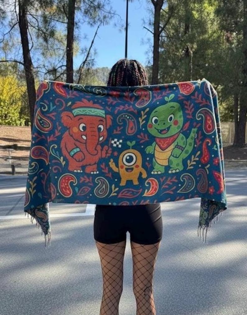 May include: A dark blue blanket with a colorful cartoon design featuring an orange elephant, a green dinosaur, and a yellow monster. The blanket has a paisley pattern and fringed edges.