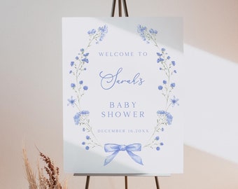 May include: A blue and white baby shower welcome sign with a floral wreath and a blue bow. The sign reads "Welcome to Sarah's Baby Shower December 16, 20XX".