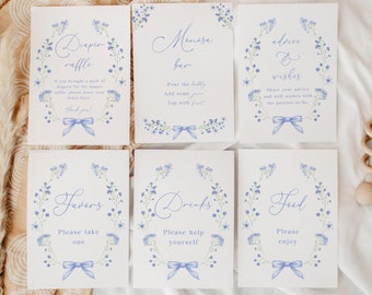 May include: Six white signs with blue floral wreaths and blue bows. The signs have text that reads "Diaper Raffle", "Mimosa Bar", "Advice & Wishes", "Favors", "Drinks", and "Food".