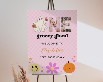 May include: A rectangular sign with a light pink checkered background featuring a cartoon ghost, a yellow smiley face, a purple flower, and an orange pumpkin. The text reads ONE, groovy ghoul, WELCOME TO, Elizabeth's, 1ST BOO-DAY.
