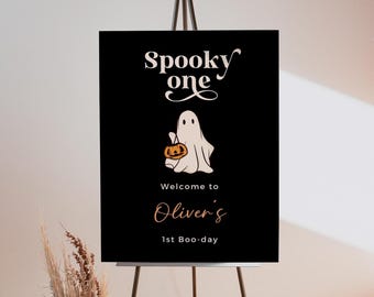 Könnte beinhalten: Schwarzes Halloween-Schild mit weißem Text "Spooky One". Ein Cartoon-Geist mit einem Kürbis und dem Text "Welcome to Oliver's 1st Boo-day". Das Schild steht auf einer schwarzen Staffelei, passend für eine Halloween-Party.