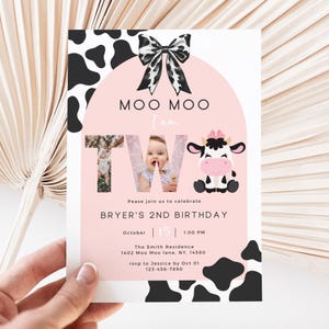 Puede incluir: Una invitación rosa y blanca con temática de vaca. La invitación dice "MOO MOO I am TW" con una foto de un bebé. También dice "BRYER'S 2ND BIRTHDAY" e incluye detalles del evento. Una mano sostiene la invitación.