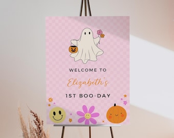 May include: A rectangular sign with a light pink checkered background featuring a white ghost holding a pumpkin bucket and flowers. The text reads WELCOME TO Elizabeth's 1ST BOO-DAY, accompanied by a yellow smiley face, a purple flower, and an orange pumpkin.