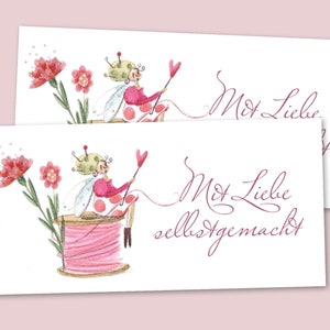 Könnte beinhalten: Eine rosa und grüne Fee mit Nadel und Faden sitzt auf einer Spule mit rosa Faden. Die Fee hält ein rosa Herz. Der Text "Mit Liebe selbstgemacht" ist in Kursivschrift unter der Fee geschrieben.