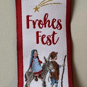 Könnte beinhalten: Besticktes weißes Stoffetikett mit roten Rändern. Der Text "Frohes Fest" in roter Schrift befindet sich über einer Illustration von zwei Figuren, die einen Esel reiten, mit einem Sternschnuppen und einem goldenen Stern. Festliche Weihnachtsdekoration.