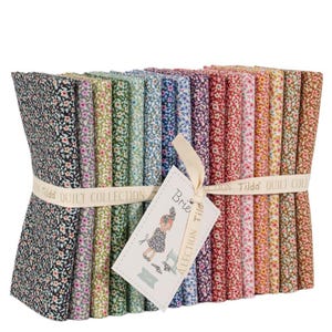 Könnte beinhalten: Ein Stapel Stoffpakete mit Blumenmuster in verschiedenen Farben, gebunden mit einem Band mit der Aufschrift "Tilda QUILT COLLECTION". Ein Etikett mit einer Zeichnung eines Mädchens und dem Text "Brie Collection Tilda" ist angebracht. Die Stoffe weisen kleine Blumenmuster auf.