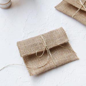 Könnte beinhalten: Ein kleiner rechteckiger Beutel aus beigefarbenem, gewebtem Jute-Stoff. Der Beutel verfügt über einen Klappenverschluss, der mit einer dünnen, metallisch goldenen Schnur gesichert ist, die in der Mitte zu einer dekorativen Schleife gebunden ist.