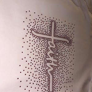 Könnte beinhalten: Weißes Langarmshirt mit Strass-Design. Das Design zeigt ein Kreuz und das Wort "Faith" in Schreibschrift, alles aus kleinen, dunkelroten Strasssteinen. Das Shirt ist aus einem weichen, weißen Stoff gefertigt.