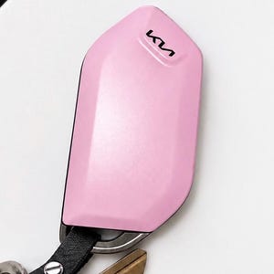 May include: A light pink protective key fob cover featuring the black Kia logo printed on the upper portion. The cover has a smooth, angular design and is attached to a black leather strap with a silver metal rivet, holding several metal keys.
