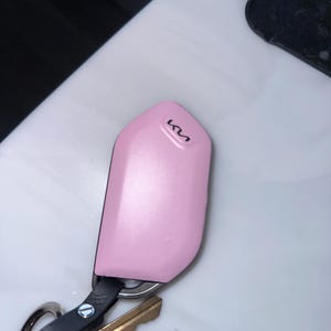 May include: A light pink protective key fob cover featuring the black Kia logo at the top. The cover is attached to a black leather strap with a silver metal screw, connected to a silver keyring holding a brass-colored key and a black object.