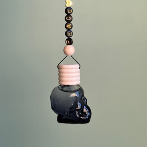 Può includere: Un profumatore per auto a forma di teschio nero con un anello di legno bianco e una corda nera con perline dorate che compongono la scritta "PEACE LOVE".