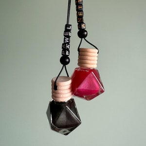 以下が含まれることがあります： 黒い紐に吊るされた、黒と金の車の芳香剤2つ。芳香剤はダイヤモンド型で、木製のトップが付いています。紐には、「PAWEL」と「ANETTA」という名前が書かれた黒いビーズが付いています。