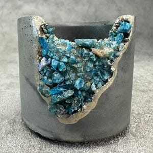 Könnte beinhalten: Grauer Beton-Pflanztopf mit goldfarbener Umrandung und einer Ansammlung leuchtend blauer Kristalle. Der zylindrische Topf hat eine raue Textur, ideal für kleine Pflanzen oder Sukkulenten.