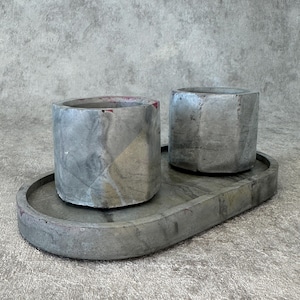 Può includere: Due vasi in cemento grigio con un design geometrico, appoggiati su un vassoio ovale in cemento grigio abbinato.