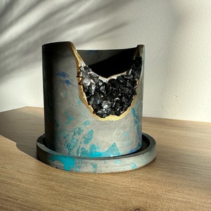 Könnte beinhalten: Ein grauer Betontopf mit goldfarbenem Rand und einem schwarzen kristallartigen Design. Der Topf hat ein blaues und graues Marmormuster.