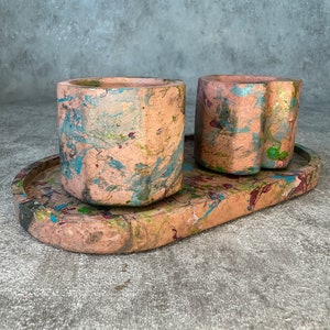 May include: Two colourful concrete planters with a matching oval tray. The planters and tray have a marbled design with shades of pink, blue, green, and brown.