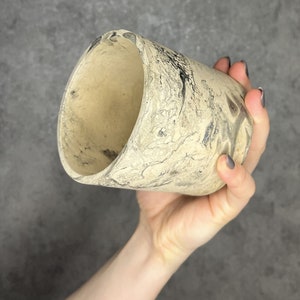 Op de afbeelding: Een kleine, beige keramische pot met een marmerpatroon. De pot wordt in een hand gehouden.