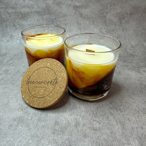 May include: Two glass candle jars with a brown and yellow wax design. The jars are sitting on a grey surface. The jars have cork lids with the text "bee concrete handmade" printed on them.